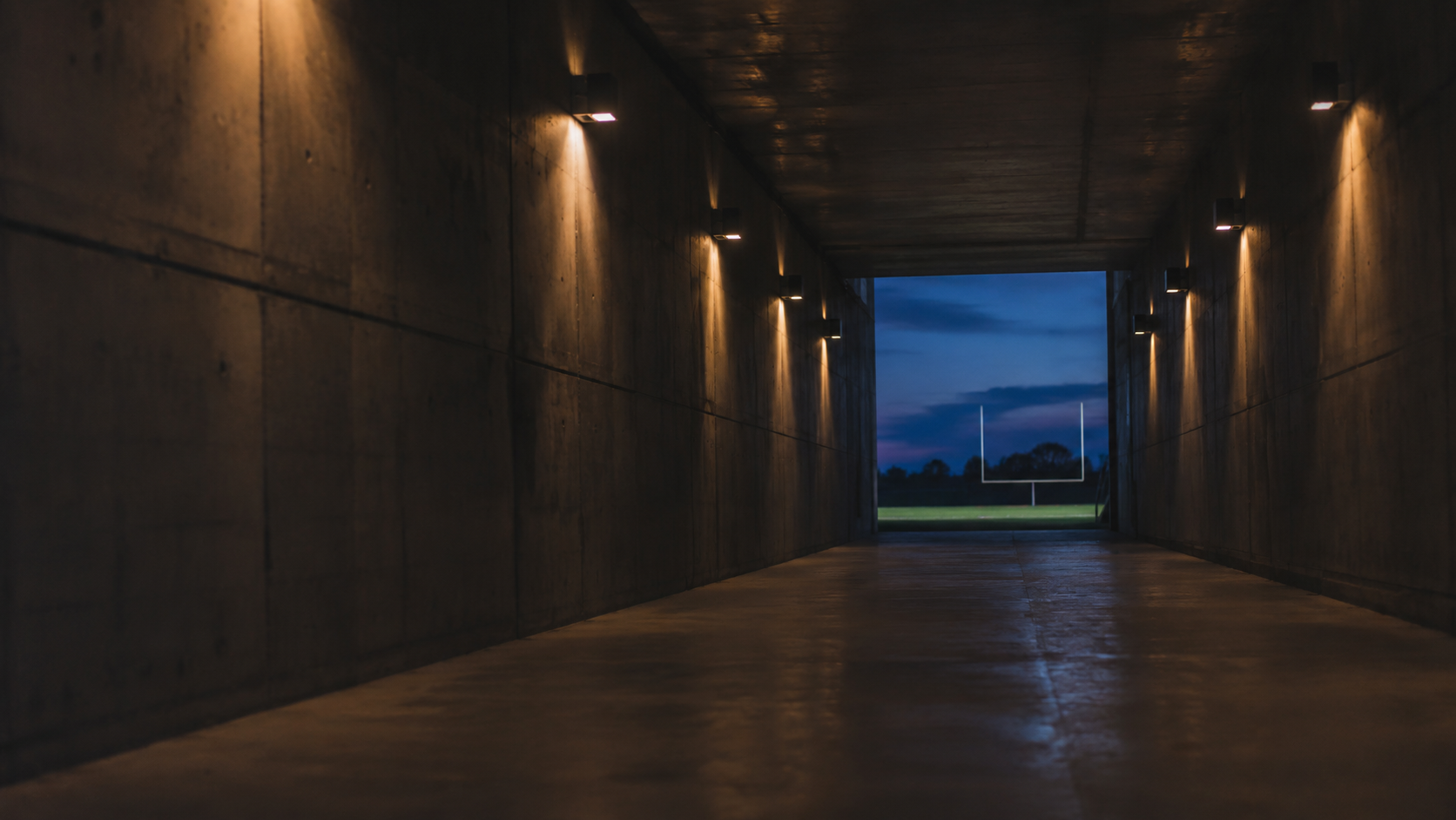 A stadium tunnel at twilight, warm tungsten lights along the walls, the field visible as a blue rectangle at the far end.